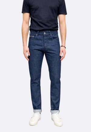 Uomo con jeans blu scuro risvoltati alle caviglie, sneakers bianche e una t-shirt nera a maniche corte, in piedi contro uno sfondo semplice.