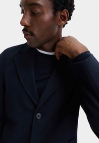Marineblauwe wollen blazer met ingesneden revers, eenknopsluiting en een gladde textuur, gedragen over een donker shirt met ronde hals.