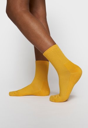 Jambes à la peau brun moyen portant des chaussettes côtelées jaunes vives jusqu'à la cheville, un pied à plat et l'autre légèrement relevé sur les orteils.