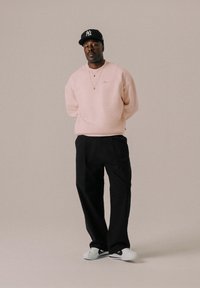 Sweat-shirt rose clair, pantalon noir à jambes larges, baskets blanches avec des accents noirs, et une casquette noire. Collier avec chaîne en or et pendentif visible.