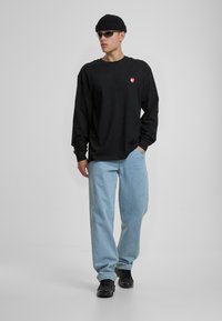 Schwarzes Langarmshirt mit kleinem rotem Logo, hellblaue lockere Jeans und schwarze Turnschuhe, ergänzt durch eine Strickmütze und Sonnenbrille.