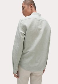Camisa de manga larga verde claro con cuello abotonado, que presenta una textura sutil y puños de un botón, combinada con pantalones de color claro.