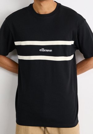 Person trägt ein schwarzes T-Shirt mit kurzen Ärmeln, zwei weißen horizontalen Streifen und weißem "ellesse"-Logo auf der Brust, kombiniert mit hellbraunen Hosen.