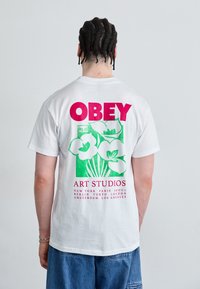 T-shirt branca com o texto em rosa intenso "OBEY" e um design floral verde nas costas, com "ART STUDIOS" e nomes de cidades impressos abaixo.