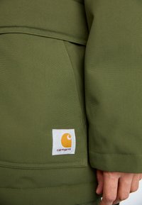 Olivenfarvet jakke med en glat tekstur, der har et hvidt mærke med et orange logo og en lomme detalje foran.