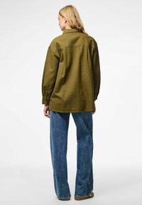 Olivgrön skjorta med avslappnad passform, knapp�ärmar och en sömm i ryggen, kombinerad med lösa blå denimjeans och ljusbruna skor.