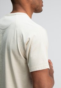 Close-up van een man in een beige shirt met korte mouwen, weergegeven van opzij en achterkant met gekruiste armen tegen een effen lichte achtergrond.