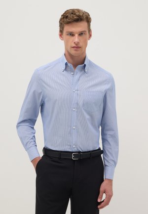Homme portant une chemise bleu clair boutonnée rentrée dans un pantalon noir avec ceinture noire, debout devant un fond clair uni.