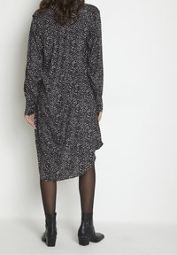 Robe noire à manches longues avec un motif tacheté blanc et gris, longueur genou, dotée d'un dos froncé et portée avec des bottines noires.