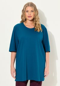 Teal übergroßes T-Shirt aus einem weichen, glatten Stoff mit rundem Ausschnitt und Ärmeln in Armlänge, kombiniert mit dunkel-violetten Hosen.