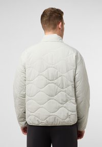 Veste matelassée gris clair avec un motif en vagues, col montant et bas élastiqué. Conçue en tissu doux et texturé.