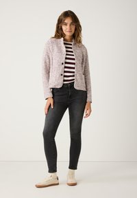 Mehrfarbige Texturjacke mit Stehkragen und Knopfverschluss, getragen über einem gestreiften Hemd, kombiniert mit schwarzen Jeans und beigen Sneakers.