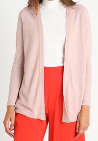 Ljusrosa öppencardigan med textilstruktur, långa ärmar och avslappnad passform, bärs över en vit ribbad polotröja och röda byxor.