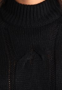 Schwarzer Strickpullover mit hohem Rippkragen, strukturierten Mustern und geflochtenen Akzenten. Das Design ist grob und eng anliegend am Hals.