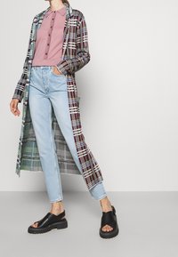 Un abrigo largo de cuadros con colores burdeos, verdes y blancos, usado sobre una camiseta tipo polo rosa y unos vaqueros de pierna recta en azul claro. Sandalias negras con suelas gruesas.