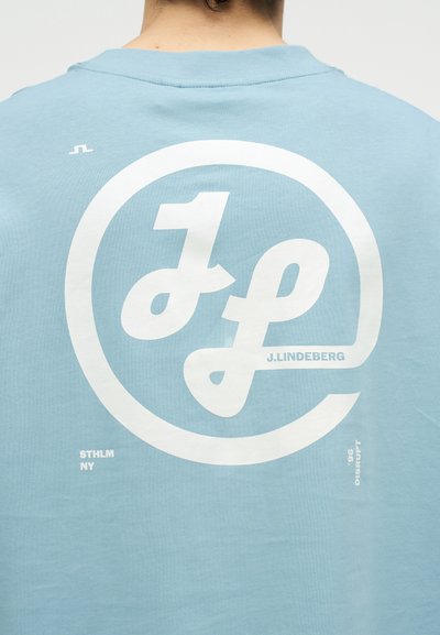 Πίσω μέρος ατόμου που φοράει ανοιχτό μπλε πουκάμισο με μεγάλο λευκό στρογγυλό λογότυπο "JL" και κείμενα "J. Lindeberg," "STHLM NY," και "Disrupt 96."