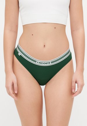 Zeleni pamukeni tange s bijelom prugastom elastičnom trakom označenom "LACOSTE", s glatkom teksturom i minimalističkim dizajnom.