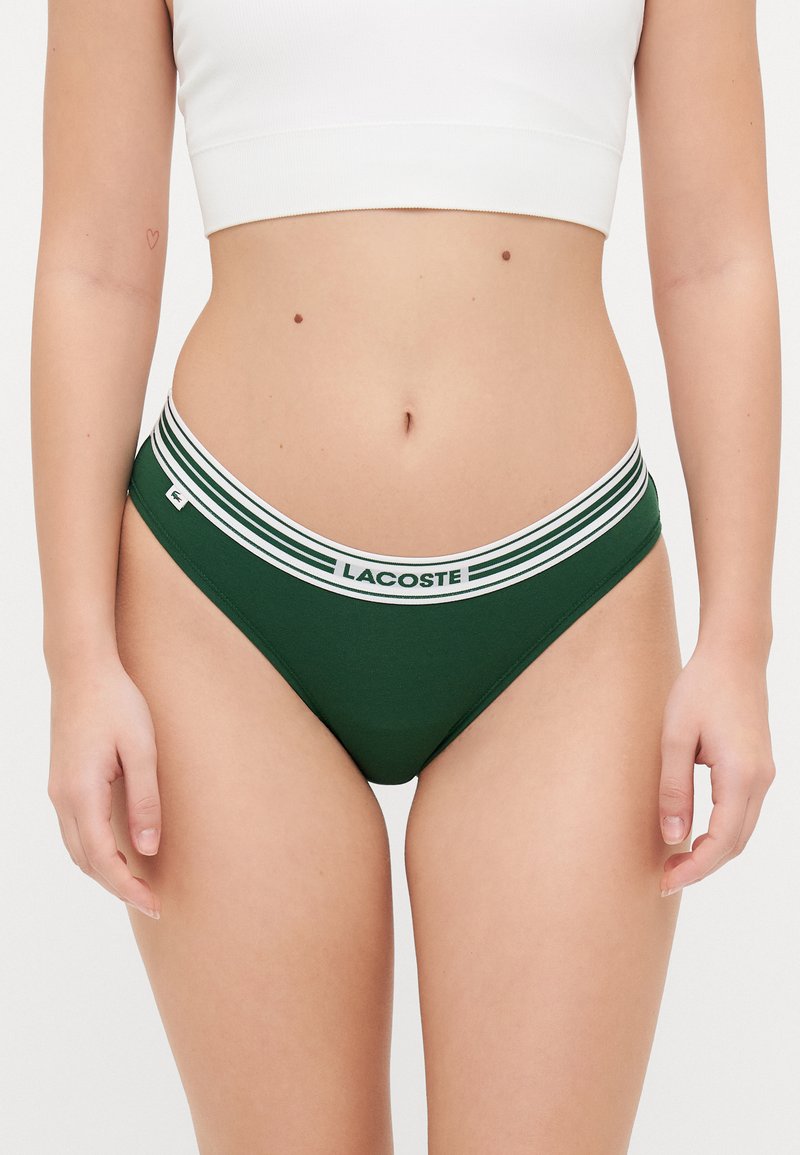 Tanga verde em algodão com uma cinta branca listada rotulada "LACOSTE", apresentando uma textura suave e um design minimalista.