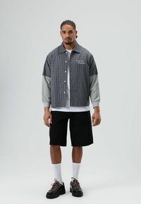 Grijs-zwart geruit overhemd met korte mouwen en geborduurde tekst, gedragen over een wit langärmelig T-shirt, gecombineerd met zwarte korte broek en sneakers.