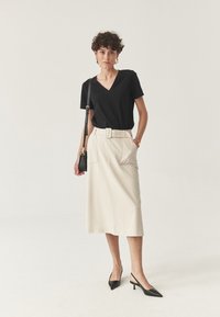 Donna che indossa un top nero a V infilato in una gonna midi beige con cintura e tasche laterali, tacchi slingback neri e una piccola borsa a spalla nera.
