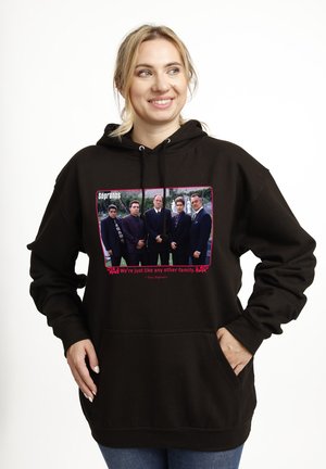 Smilende kvinde iført en sort hættetrøje med et foto af Sopranos rollebesætning og citatet "Vi er ligesom alle andre familier."