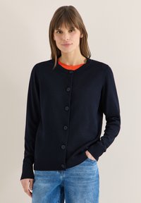 Femme portant un cardigan noir boutonné sur une chemise orange et un jean bleu, debout devant un fond clair uni.
