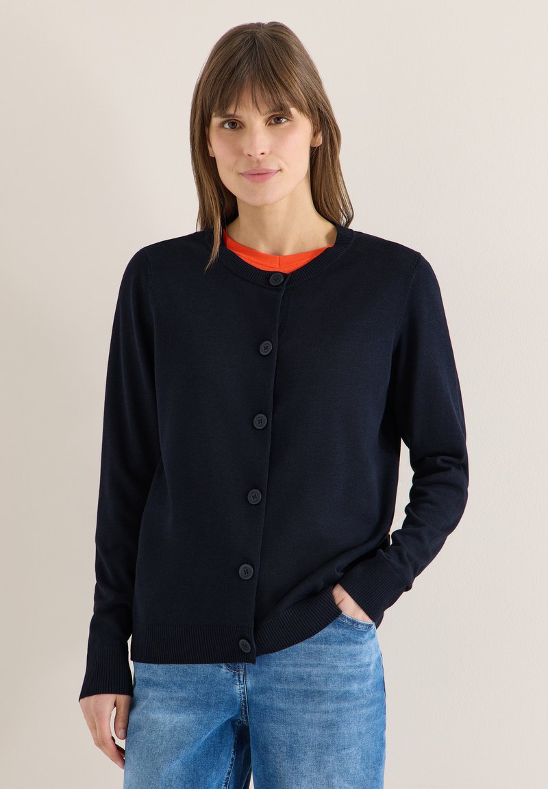 Femme portant un cardigan noir boutonné sur une chemise orange et un jean bleu, debout devant un fond clair uni.