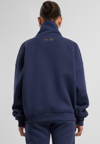 Felpa oversize blu navy con colletto alto, polsini e orlo a costine. Presenta un logo dorato sul retro. Realizzata in materiale morbido e testurizzato.