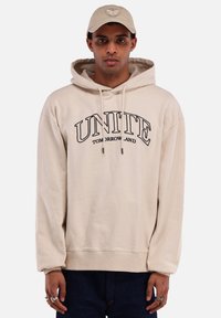 Beige hoodie met een groot zwart "UNITE TOMORROWLAND" logo. Voorzien van een trekkoord in de capuchon en geribbelde mouwen. Draagt bij donkere jeans en een beige pet.