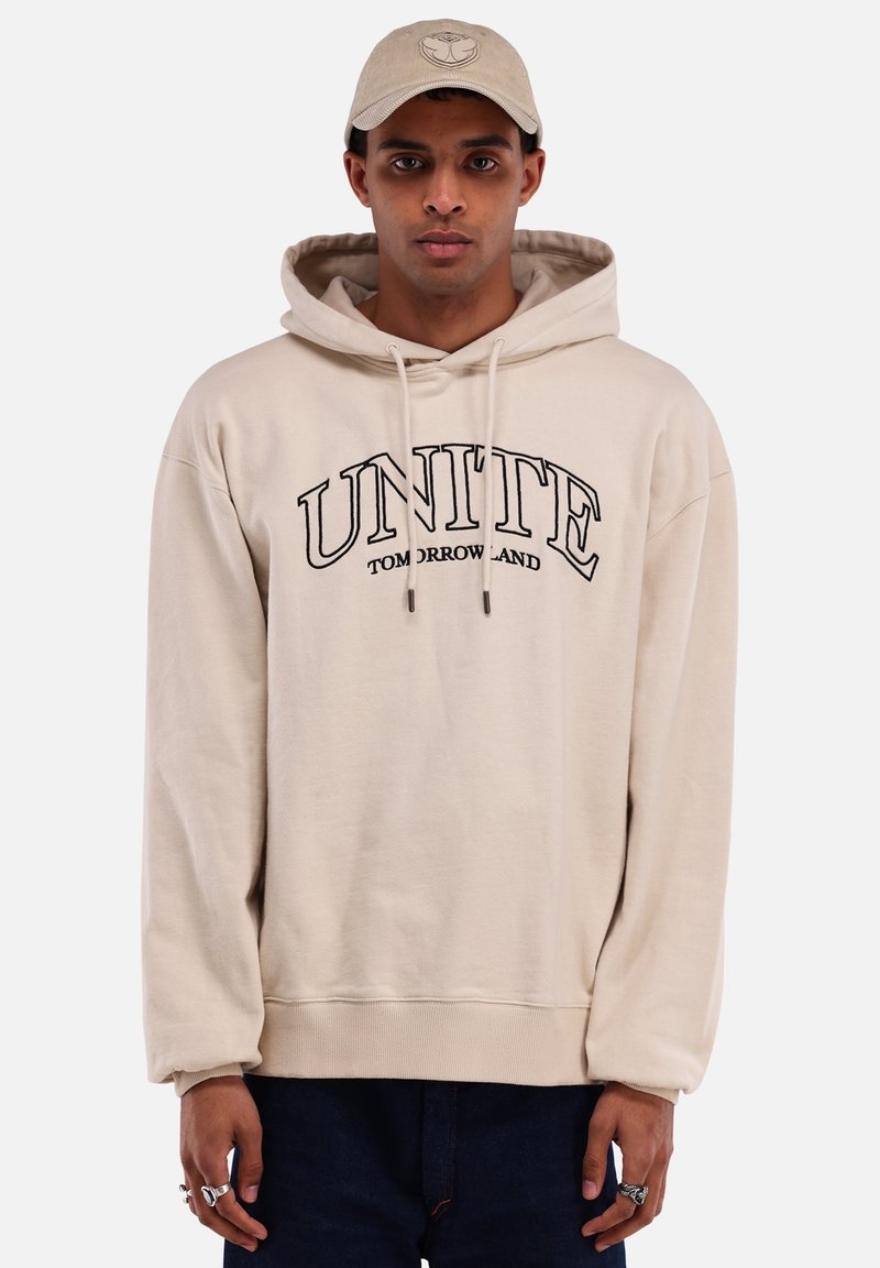 Beige hoodie met een groot zwart "UNITE TOMORROWLAND" logo. Voorzien van een trekkoord in de capuchon en geribbelde mouwen. Draagt bij donkere jeans en een beige pet.