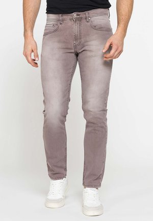 Uomo che indossa jeans grigi slim-fit sbiaditi e sneakers bianchi, in piedi davanti a uno sfondo chiaro uniforme.