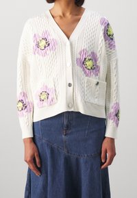 Kobieta ubrana w białą kardigan o splocie warkoczowym z purpurowymi i żółtymi kwiatowymi wzorami, z przodem zapinanym na guziki, kieszeniami i niebieską spódnicą dżinsową.