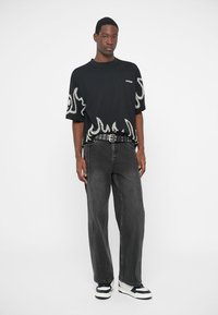 Camiseta negra de gran tamaño con patrón de llamas blancas en las mangas y el dobladillo inferior, combinada con pantalones grises holgados y zapatillas en blanco y negro.