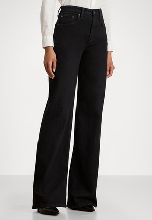 Person iført sorte jeans med højtaljede, vide ben, hvid langærmet skjorte og sorte spidse sko, stående foran en ensfarvet baggrund.