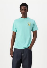 T-shirt de algodão verde menta com gola redonda e mangas curtas. Apresenta uma estampa gráfica multicolorida de flores e uma coroa no lado esquerdo do peito.