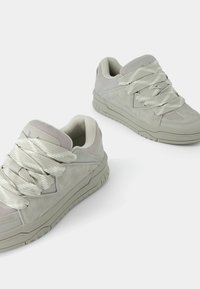 Jasnoszare trampki wykonane z zamszu i siateczki, z grubymi szarymi sznurowadłami, wyściełanym kołnierzem i teksturowanymi gumowymi podeszwami o sportowym designie.