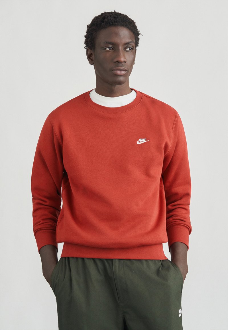 Sweatshirt vermelha de algodão com decote redondo e mangas longas, apresentando um pequeno logótipo branco da Nike no lado esquerdo do peito. Combinada com calças verdes.