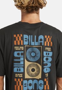 Osoba z krótkimi kręconymi włosami, nosząca czarną koszulkę z niebieskim, beżowym i pomarańczowym geometrycznym wzorem oraz tekstem na plecach.