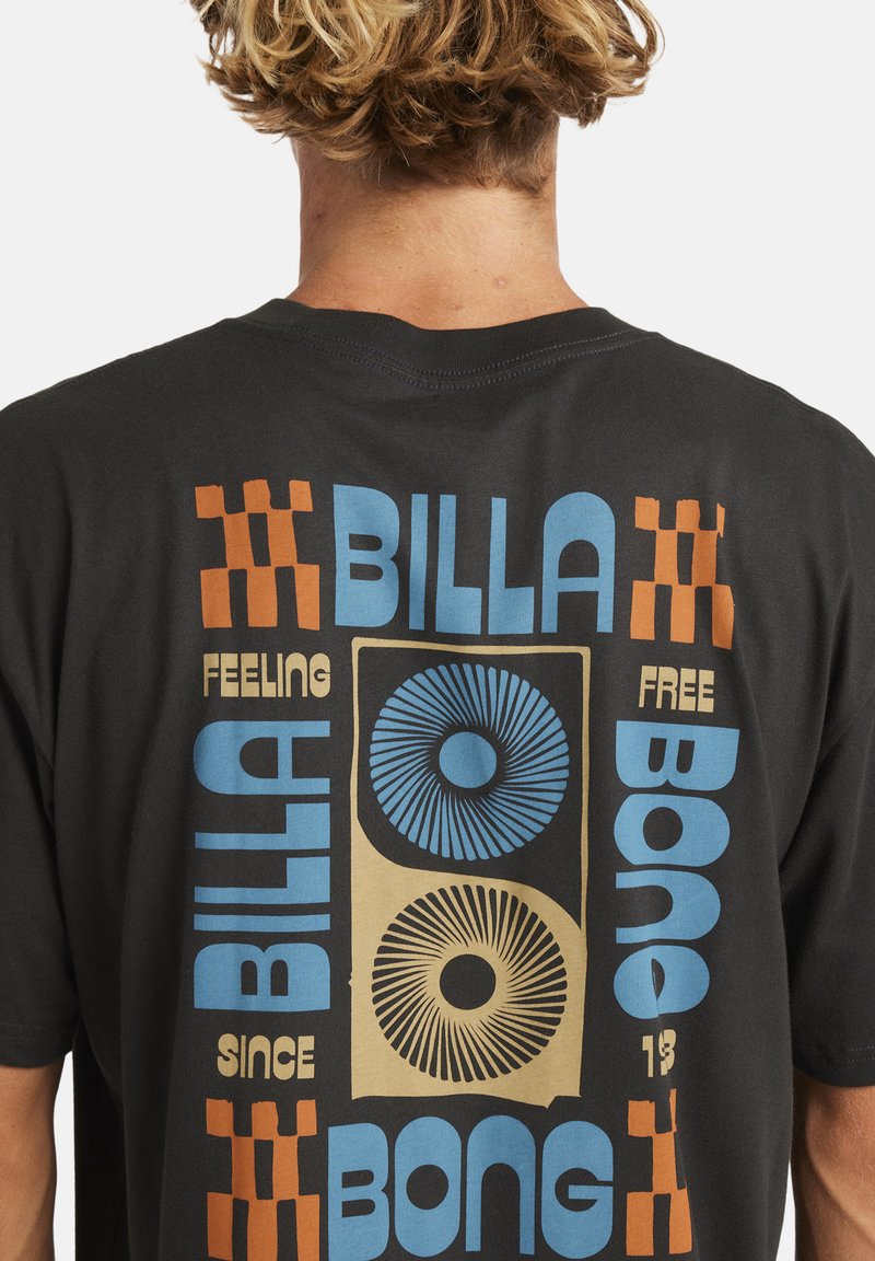 Osoba z krótkimi kręconymi włosami, nosząca czarną koszulkę z niebieskim, beżowym i pomarańczowym geometrycznym wzorem oraz tekstem na plecach.