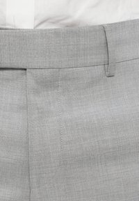 Calças cinza claro com um tecido texturizado e tecido. Apresentam uma frente lisa, silhueta justa e detalhes sutis costurados ao longo das costuras.