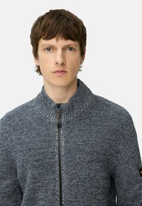 Grijze gebreide zip-up trui met hoge kraag, textuurpatroon en zwarte ritssluiting. Heeft een patch op de linkermouw.