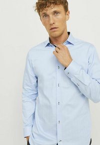 Chemise bleu clair à manches longues, avec des poignets boutonnés et un col noir contrastant. Présente des boutons décoratifs le long de la patte de boutonnage.