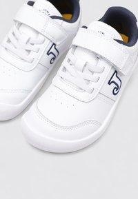 Zapatillas deportivas blancas con un parte superior sintética lisa, lengüeta acolchada, correa de velcro ajustable y logo en azul marino.