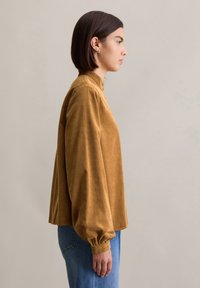 Blouse en velours marron avec col haut, manches ballon, et coupe décontractée, associée à un jean bleu clair.