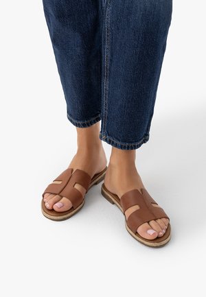 Stopy w brązowych skórzanych sandałach typu slip-on z odkrytymi palcami, zestawione z ciemnoniebieskimi dżinsami skróconymi nad kostkami.