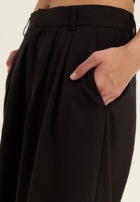 Mână odihnindu-se în buzunarul pantalonilor plisati negri cu talie înaltă, pe un fundal deschis, concentrându-se pe talie și zona buzunarului.