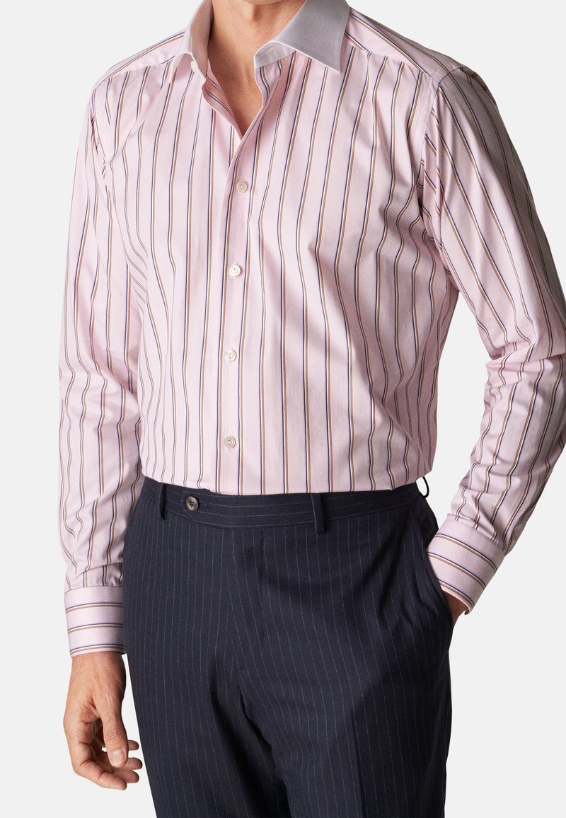 Homme portant une chemise à rayures rose clair avec un col blanc et un pantalon sombre à fines rayures, une main dans la poche.