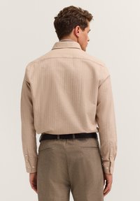 Mann, der sich abwendet und ein beiges langärmliges Hemd, das in braune Hose mit schwarzem Gürtel gesteckt ist, vor einem einfarbigen Hintergrund trägt.
