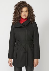 Cappotto nero in misto lana con colletto alto, vita cintrata e tasche laterali con zip. Presenta dettagli testurizzati sulle spalle e maniche dritte.