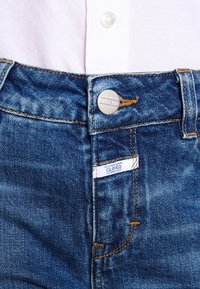 Jean en denim d'un bleu moyen avec des coutures jaunes. Il présente un bouton en métal et une étiquette sur la ceinture, affichant "CLOSED".